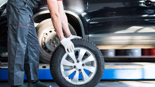 Mechanic in gloves rolling a car tire toward a lifted vehicle.